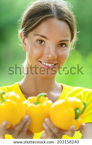 Portrait of dark-haired smiling beautiful young woman in yellow blouse with bell pepper, against green of summer park.