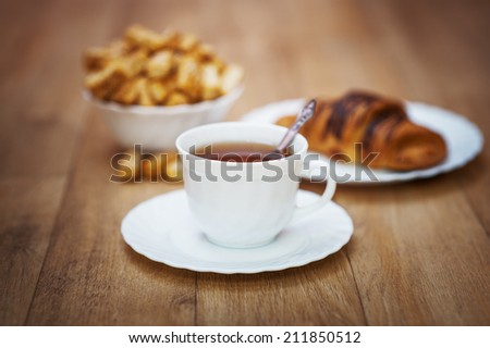 Close-up on wooden kitchen table, black strong coffee, topped with sesame seeds small cookies and chocolate croissant (all products in a beautiful snow-white ware