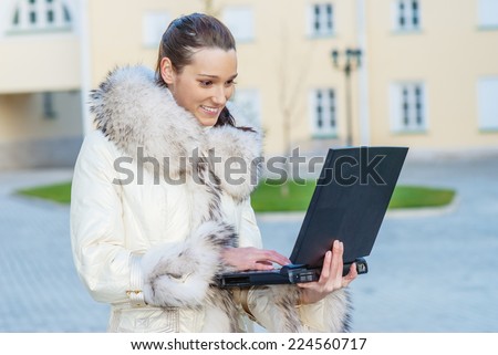 Young smiling beautiful woman in white coat with fur collar sitting on bench and holding laptop on your lap.
