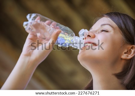 Portrait of young beautiful woman with bare shoulders drinks from bottle of cold water, on green background summer nature.