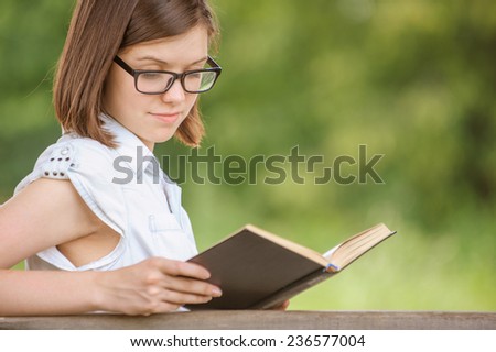 Portrait of cute brunette girl wearing goggles and white chemise reading exciting book at summer green park.