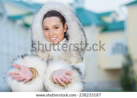 Young smiling beautiful woman in white coat with fur collar on background of buildings with green roofs.
