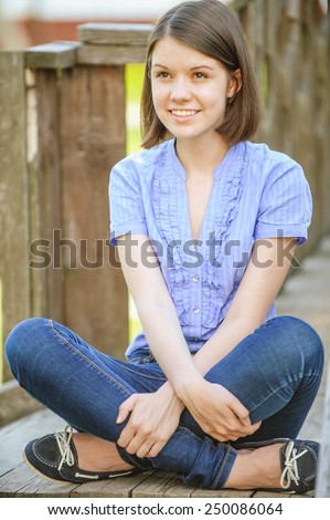 Young beautiful dark-haired smiling woman wearing blue blouse and jeans sitting on wooden staircase at summer park.