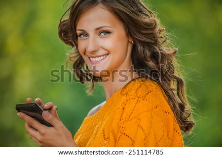 Beautiful young woman close-up in orange sweater is typing with stylus on device, against green of summer park.