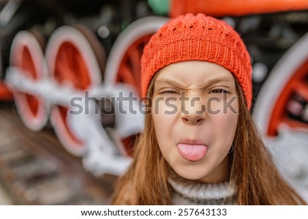 Little beautiful girl in a red hat shows tongue, against the background of an old steam locomotive.