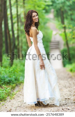 Young beautiful bride in white wedding dress with bare feet standing on bridge over stream in summer park.