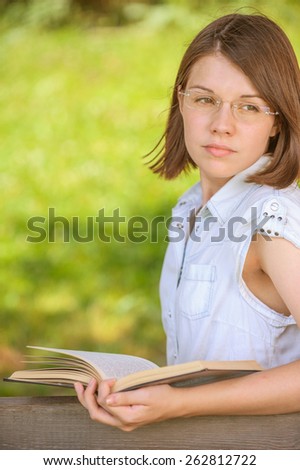 Portrait of young attractive brunette woman wearing glasses, reading book, sitting at summer green park