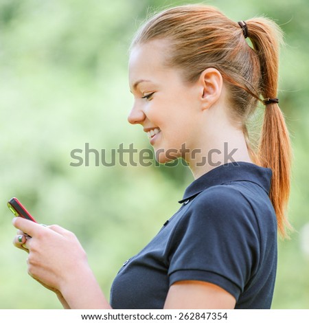 Beautiful smiling young woman profile in dark blouse talking on mobile phone, against green of summer park.