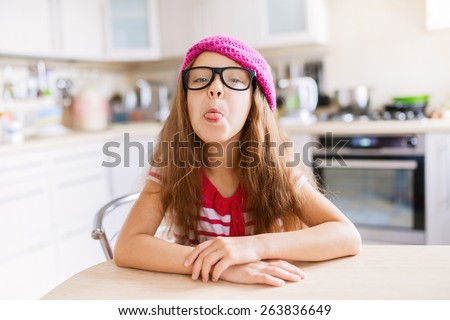 Beautiful little girl in a red cap sits at a table in the kitchen and shows tongue.
