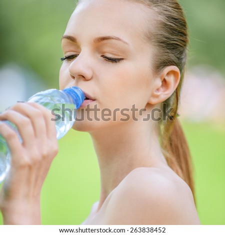 Beautiful young woman drinks water from bottle, against background of summer green park.