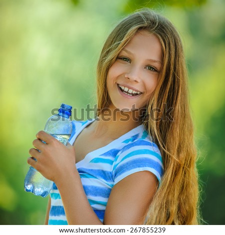 Beautiful teenage girl in blue blouse with water bottle, against green of summer park.