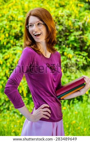Beautiful red-haired smiling young woman in a purple blouse with books, against green summer city park.