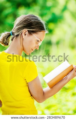 Dark-haired smiling beautiful young woman in yellow blouse and glasses reading books profile, against green of summer park.