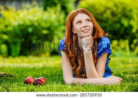 Beautiful red-haired smiling young woman in a blue blouse lying on the grass, on a background of green summer city park.