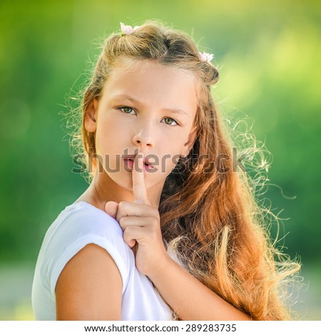 Beautiful teenage girl in white blouse raised index finger to her lips, against green of summer park.