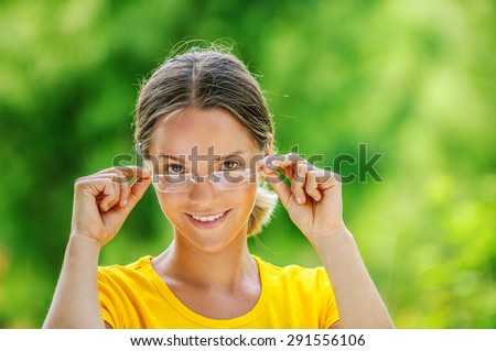 Dark-haired smiling beautiful young woman in yellow blouse and glasses, against green of summer park.