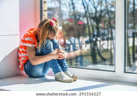 Little girl in orange blouse sits on a window sill, crying.