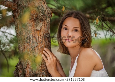 Dark-haired smiling beautiful young woman in white blouse near trunk tree, against green of summer park.