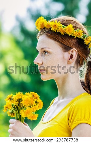 Beautiful red-haired smiling young woman in yellow blouse with a wreath and a bouquet of dandelions on a green background of summer city park.