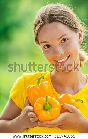 Portrait of dark-haired smiling beautiful young woman in yellow blouse with bell pepper, against green of summer park.