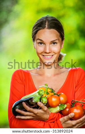 Portrait of dark-haired smiling beautiful young woman in red blouse with tomatoes, eggplants, peppers, against green of summer park.
