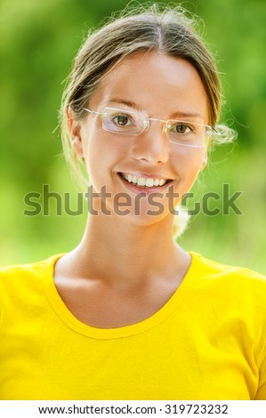 Dark-haired smiling beautiful young woman in yellow blouse and glasses, against green of summer park.