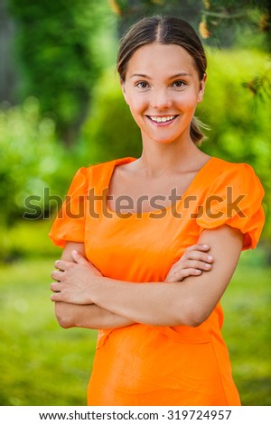Portrait of dark-haired smiling beautiful young woman in orange blouse, against green of summer park.