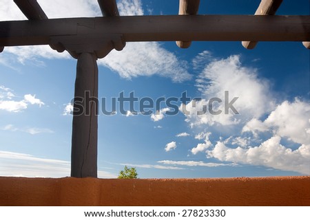Southwestern architecture. Beams and earth colored wall against blue sky, one little tree showing up behind the wall. New Mexico