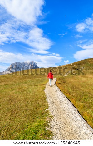Young woman tourist walking alpine trail with view of Sassolungo mountain range, Passo Gardena, Sudtirol, Dolomiti, Italy