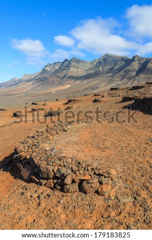 Walls built of lava stones protect wine plants from wind on sunny slopes of mountains near Cofete beach, Fuerteventura, Canary Islands, Spain
