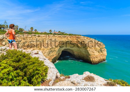 Young woman tourist standing on cliff rock and looking at sea cave on coast of Portugal, Algarve region
