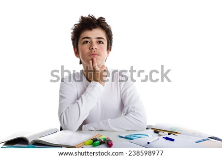 Caucasian young boy sits in front of a desk covered by his homework: he meditates with questioning eyes on blank book and holds his chin with right hand