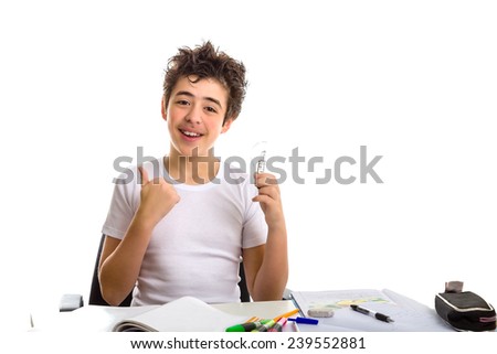Boy is doing homework: sitting in front of a desk covered by books,copybooks and pencils, he holds a lightbulb with his left hand and makes succcess gesture with right hand
