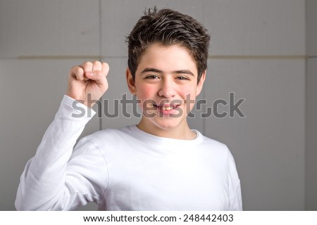 Latin smooth-skinned boy in a white long sleeved t-shirt smiles making a gesture of power raising his right fist  on industrial background