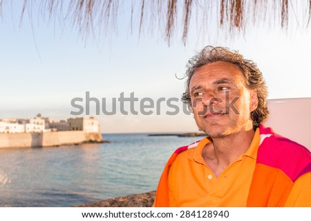 Handsome tanned middle-aged man with salt pepper hair with red and orange sweater and yellow polo shirt in Italian outdoors: he is relaxed under palm while antique buildings are facing on seaside