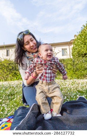 Cute 6 months old baby with Light brown hair in red checkered shirt and beige pants is embraced and held by his smiling Hispanic mummy in a green city park