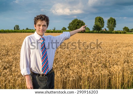 Handsome Caucasian boy wearing a white shirt and a regimental tie with red, fuchsia, orange, blue, indigo and white stripes is pointing  on a field of golden wheat ears at sunset