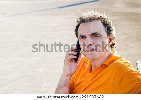 Classy senior sportsman with three-day beard and salt and pepper hair wearing an orange polo shirt while he is sitting on a bench on the pier and talking on a mobile phone