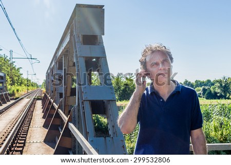 Caucasian 40 years old man, elegant and sporty dressing a blue polo shirt,  is talking on the mobile phone next to railroad tracks in the Italian countryside in summer