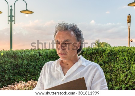Classy senior sportsman with three-day beard and salt and pepper hair wearing a white shirt while reding the restaurant menu