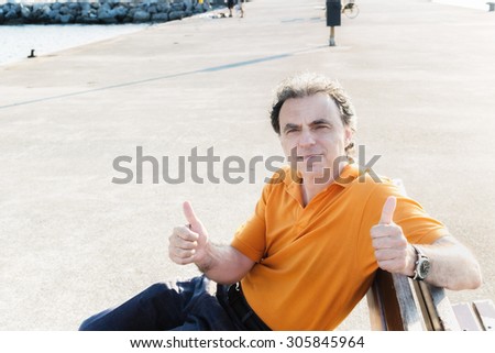 Classy  40 years old sportsman with three-day beard and salt and pepper hair wearing an orange polo shirt while he is sitting on a bench on the pier and showing thumbs up