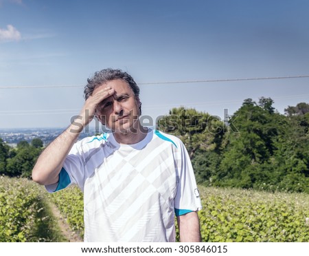 Handsome 40 years old man with salt pepper hair dressed with sports shirt is holding his head in the cultivated fields of Italian countryside: he seems to have headache