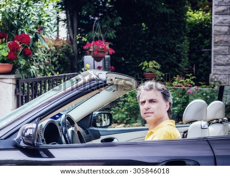 Side view of classy  40 years old sportsman with three-day beard and salt and pepper hair wearing a yellow polo shirt while he is driving a dark brown car in residential neighborhood