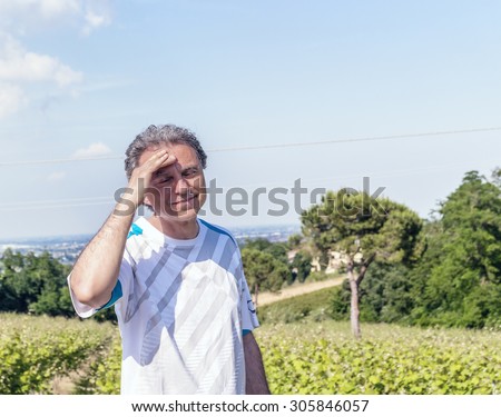 Handsome 40 years old man with salt pepper hair dressed with sports shirt is holding his head in the cultivated fields of Italian countryside: he seems to have headache