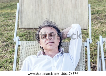 summer retreat away from the stress - charming Caucasian middle-aged man with gray hair and white shirt resting on a deck chair in the countryside
