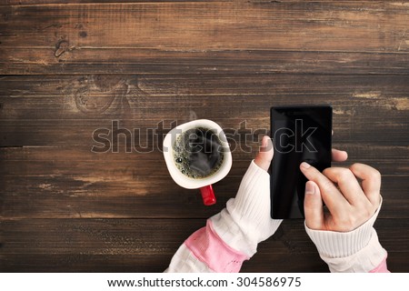 Above view of female hand holding smart phone with hot cup of coffee on wood table. Photo in vintage color image style.