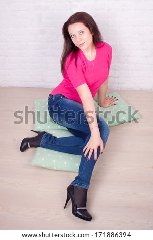 Girl in a pink blouse and blue jeans. Brunette. On the background of a brick white wall. Parquet floor. Sitting on pillows