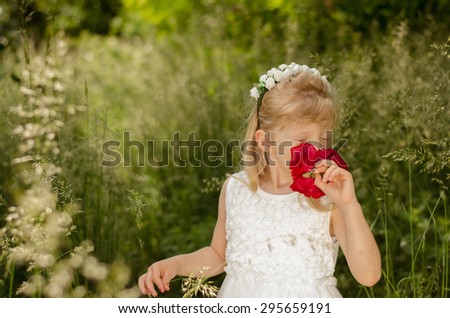 adorable blond little girl with flower headband smelling red rose in green meadow