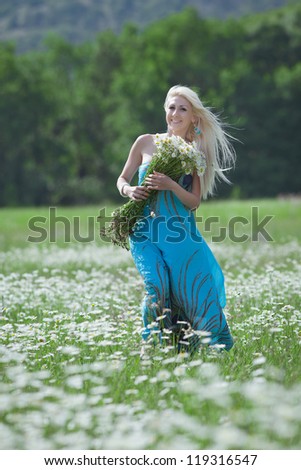 Attractive blonde in chamomile field. Young woman with bouquet of flowers walking through chamomile field