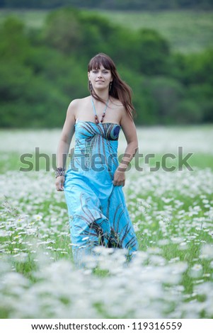Attractive brunette in chamomile field. Young woman in blue sundress walking through chamomile field